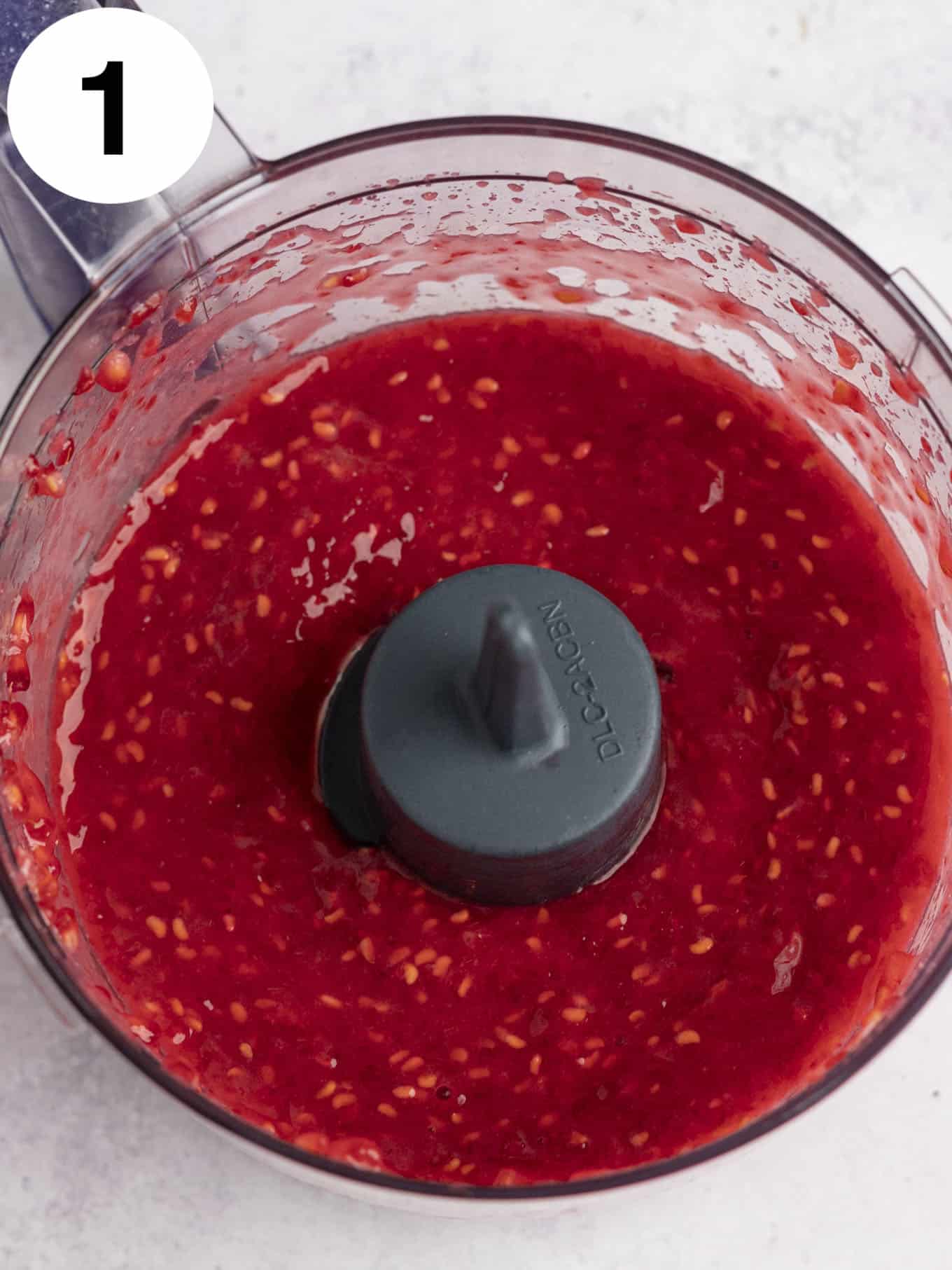 An overhead view of pureed raspberries in the bowl of a food processor. 