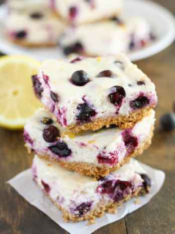 A stack of three lemon blueberry cheesecake bars. A lemon half and additional bars rest in the background.