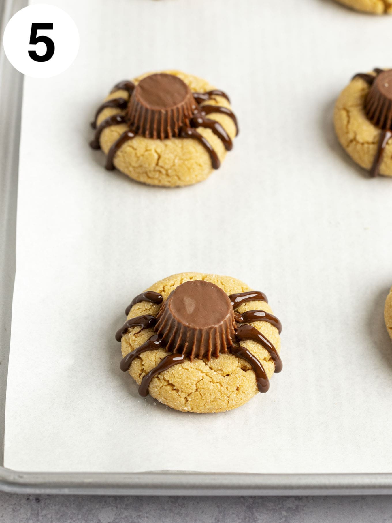 A process shot showing peanut butter cup cookies with spider legs piped onto them with melted chocolate. 