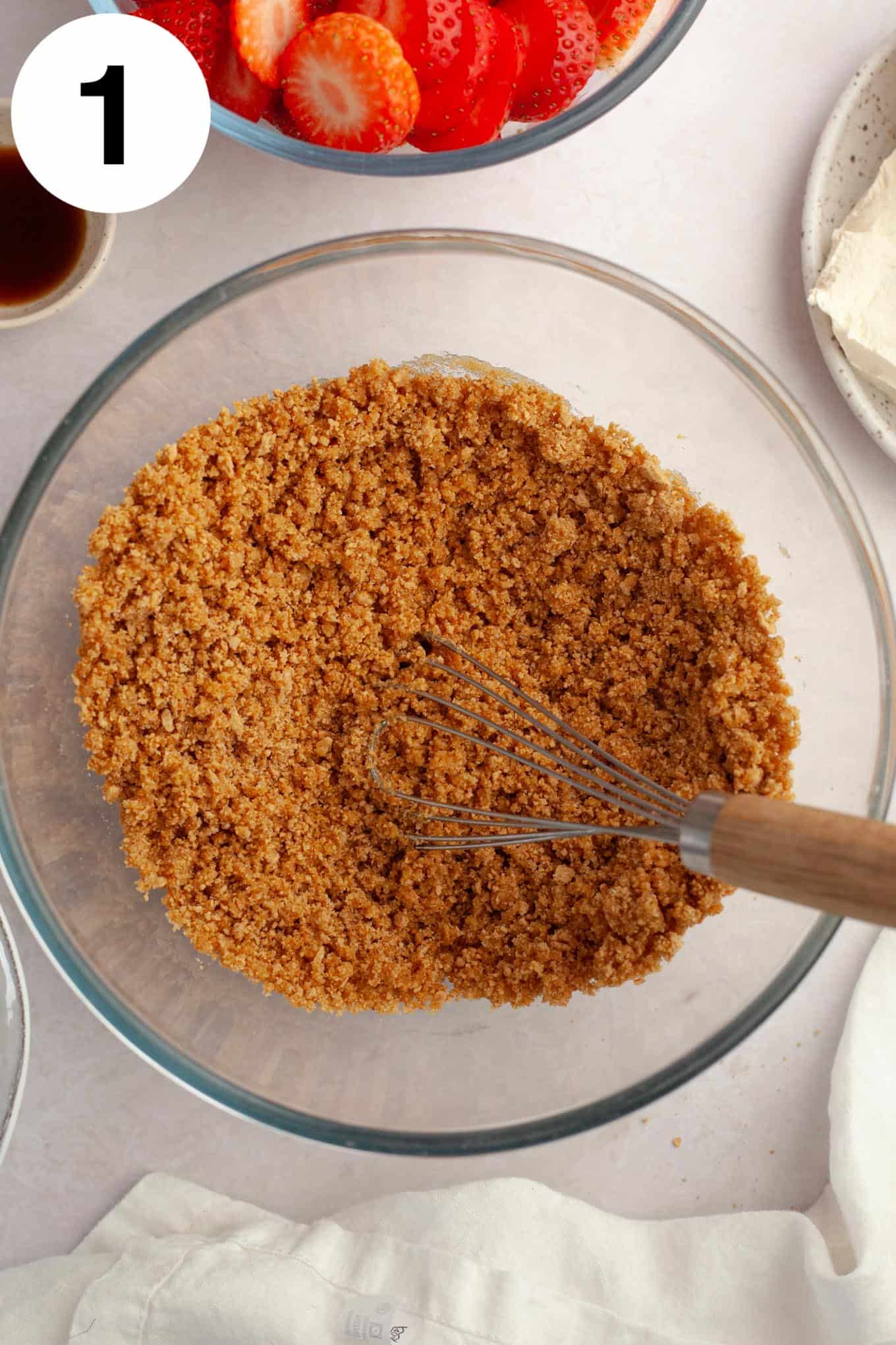 An overhead view of graham cracker crumbs in a glass mixing bowl, with a whisk. 