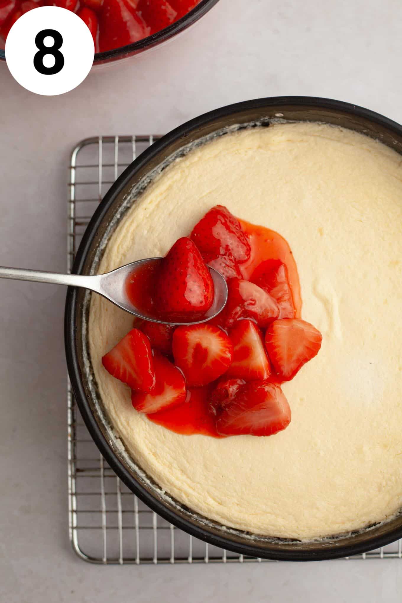 An overhead view of strawberry topping being spooned over a baked cheesecake. 