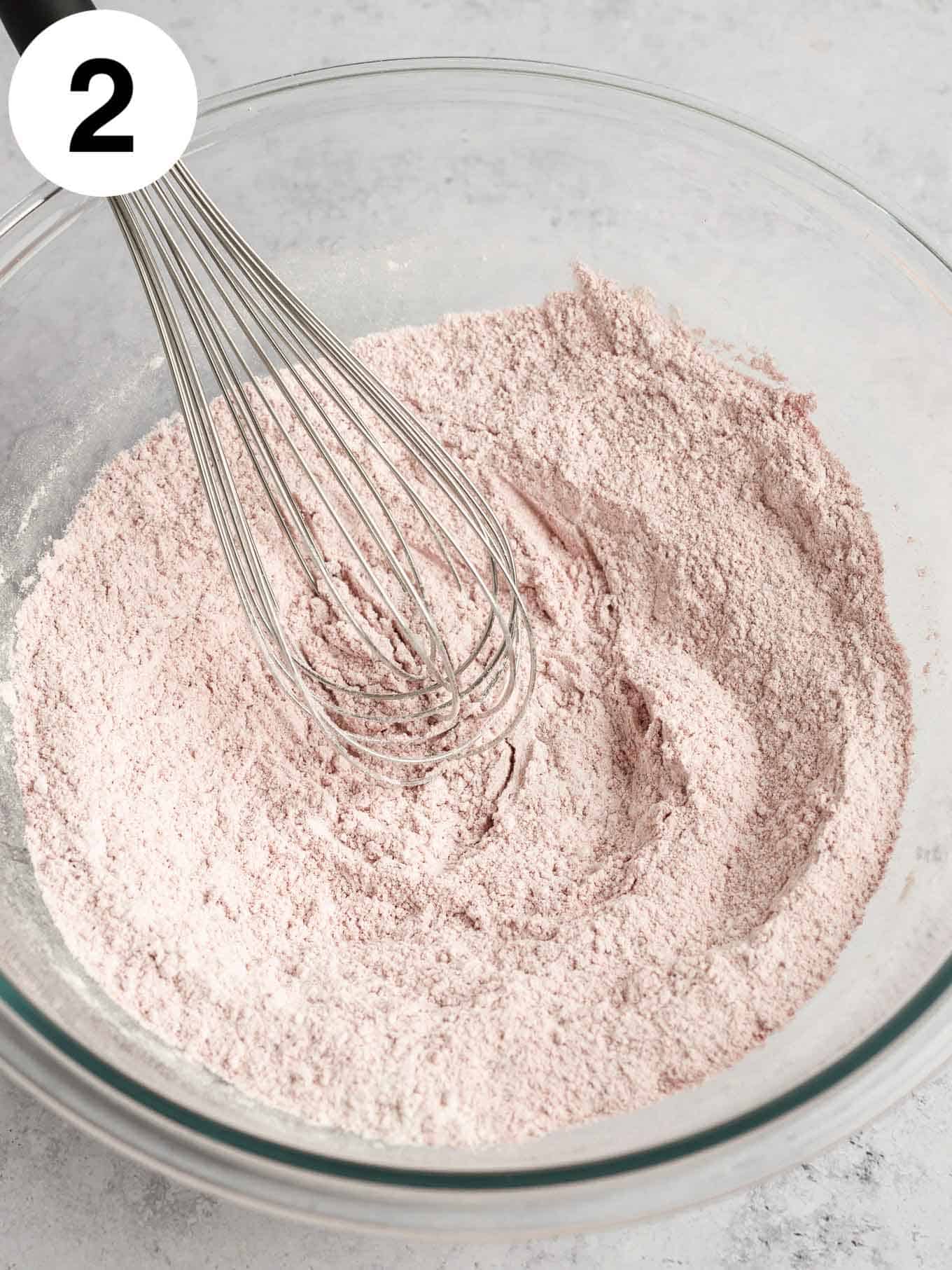 An overhead view of the dry ingredients in a glass mixing bowl, with a whisk. 
