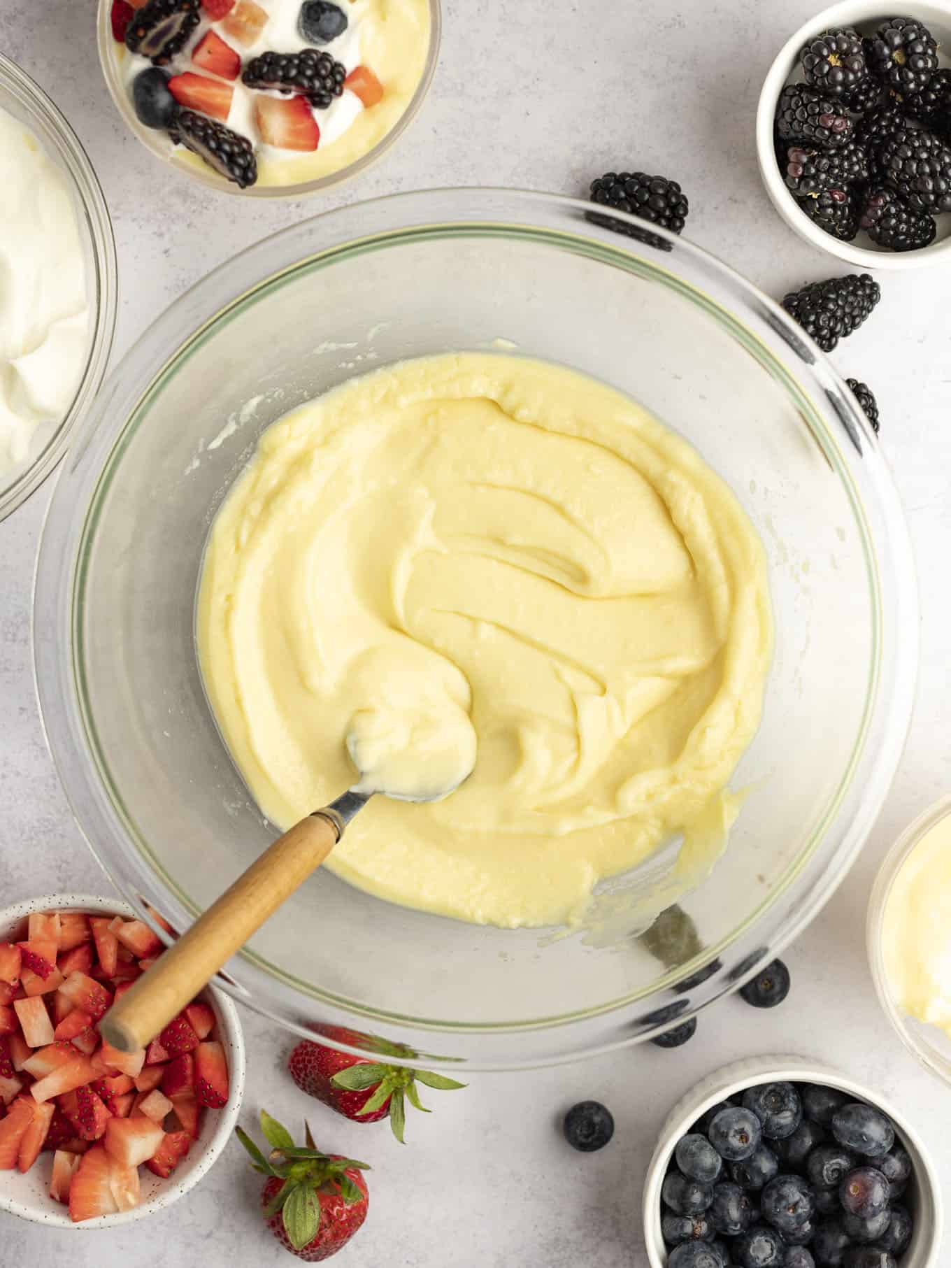 An overhead view of homemade vanilla pudding in a glass bowl, surrounded by dishes of fresh berries. 