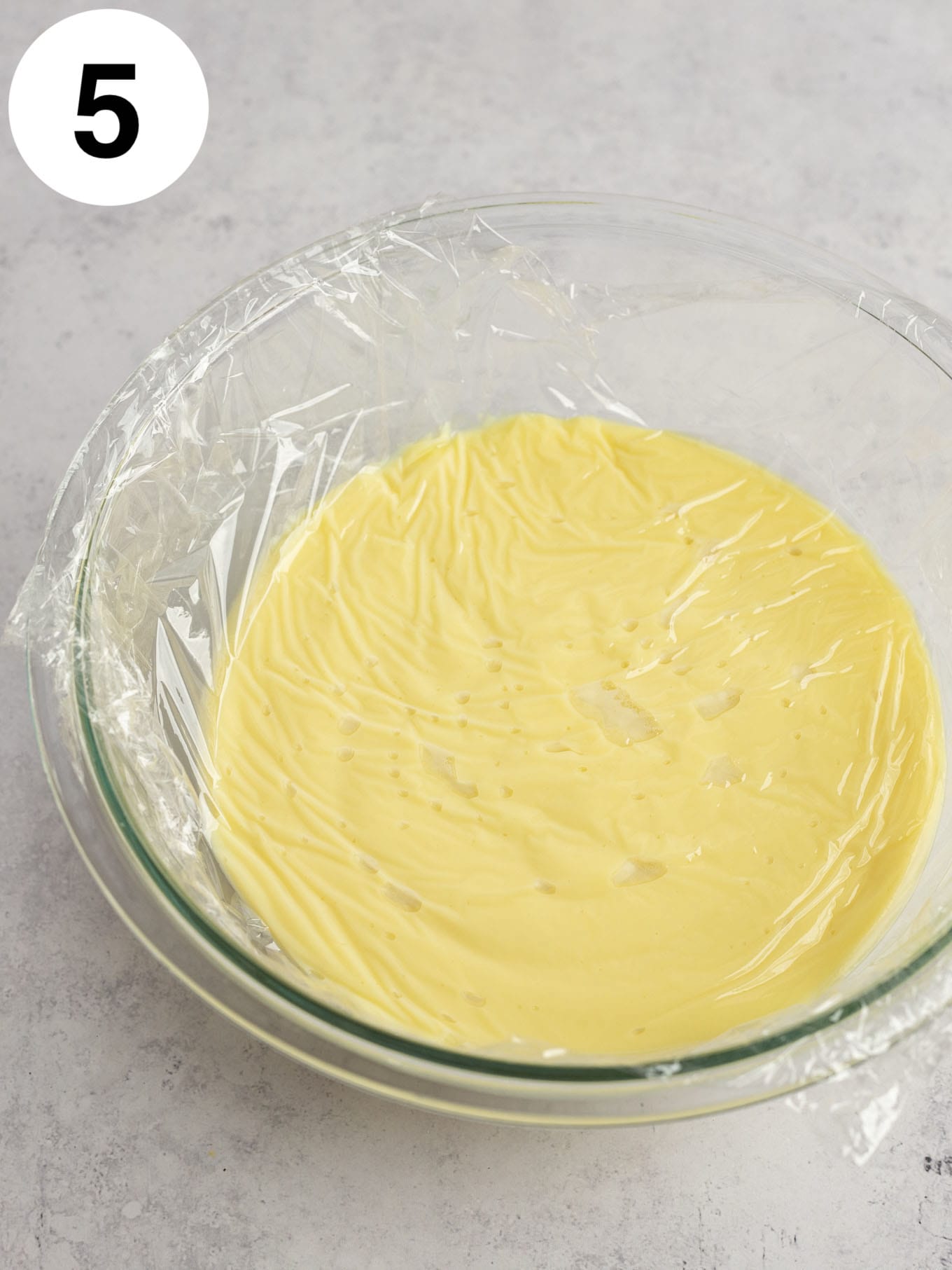 Vanilla pudding in a glass bowl, with a piece of plastic wrap pressed on top. 