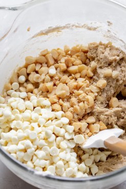 A glass mixing bowl filled with cookie dough topped with white chocolate chips and nuts being mixed together with a rubber spatula.