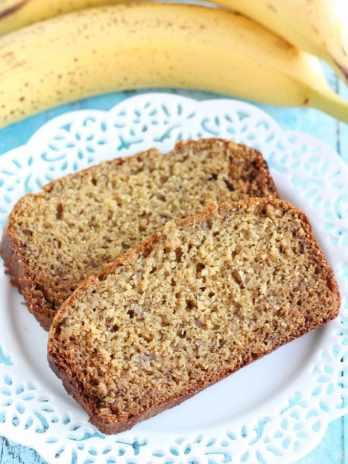 Two slices of whole wheat banana bread on a white plate. Two bananas rest in the background behind the plate.