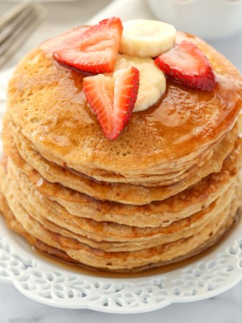 A stack of whole wheat pancakes topped with strawberry, banana slices, and maple syrup.